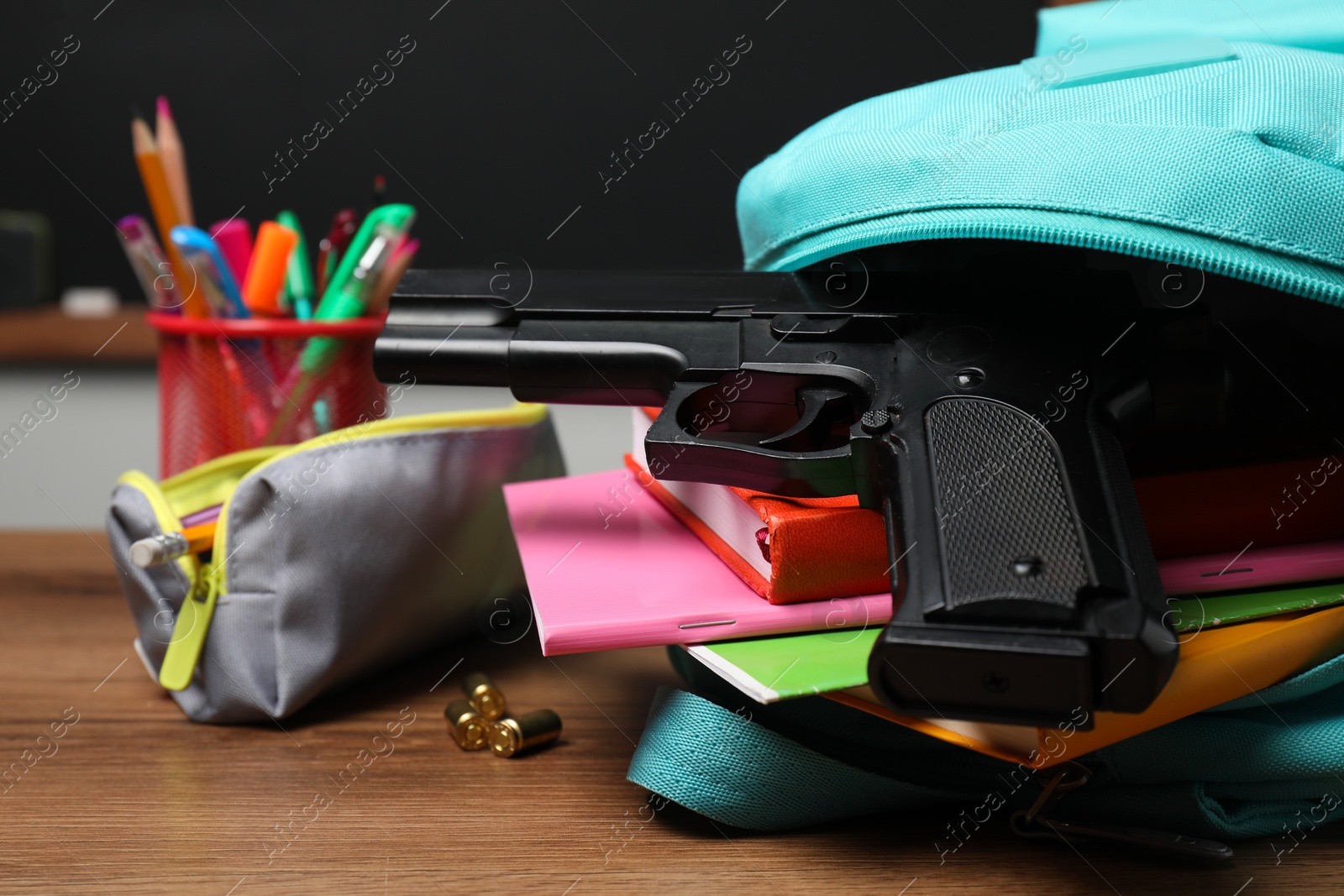 Photo of Gun, bullets and school stationery on wooden table near blackboard indoors, closeup