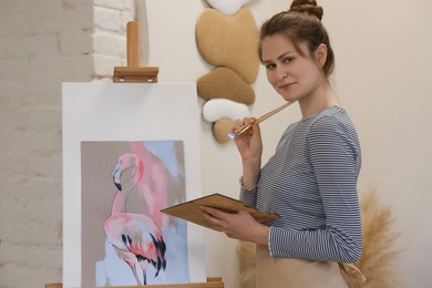 Portrait of woman with brush and palette near her artwork in drawing studio