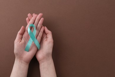 Woman holding turquoise awareness ribbon on brown background, top view. Space for text