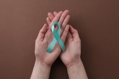 Woman holding turquoise awareness ribbon on brown background, top view