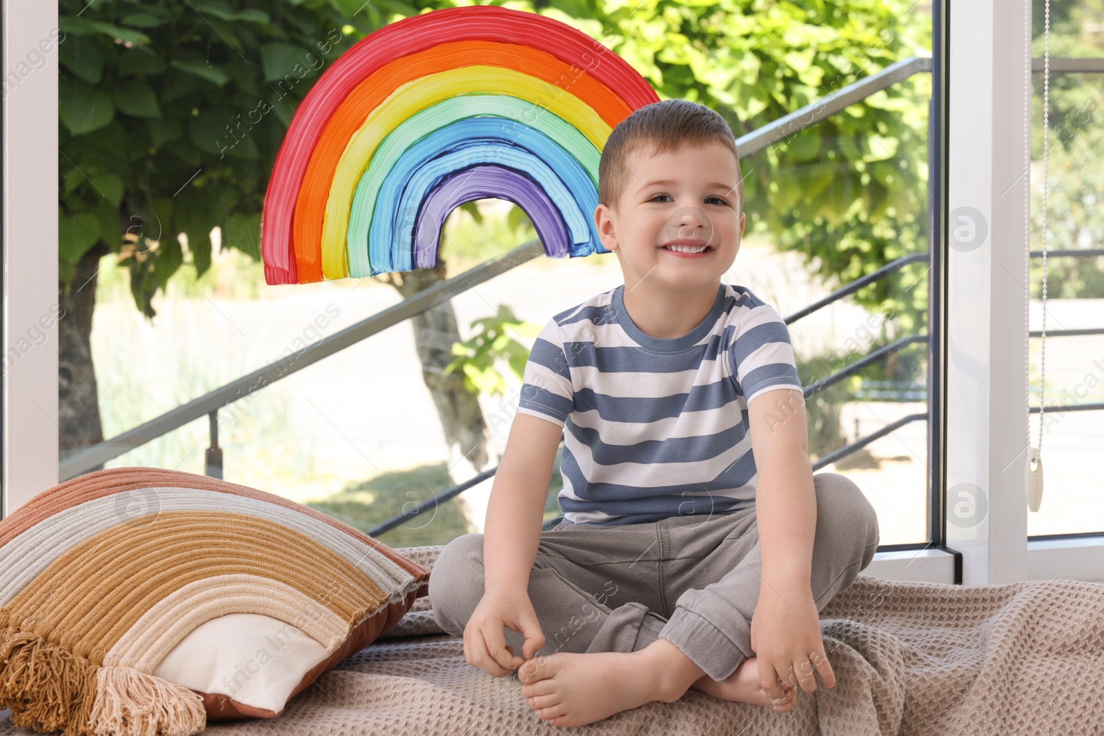 Photo of Happy little boy near rainbow painting on window indoors