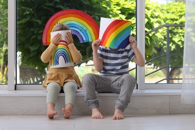Children covering faces with pictures of rainbow near window indoors