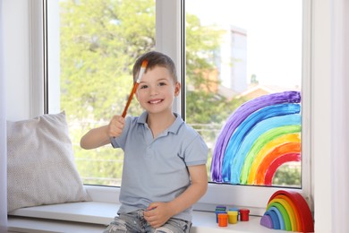 Photo of Little boy with brush near rainbow painting on window indoors