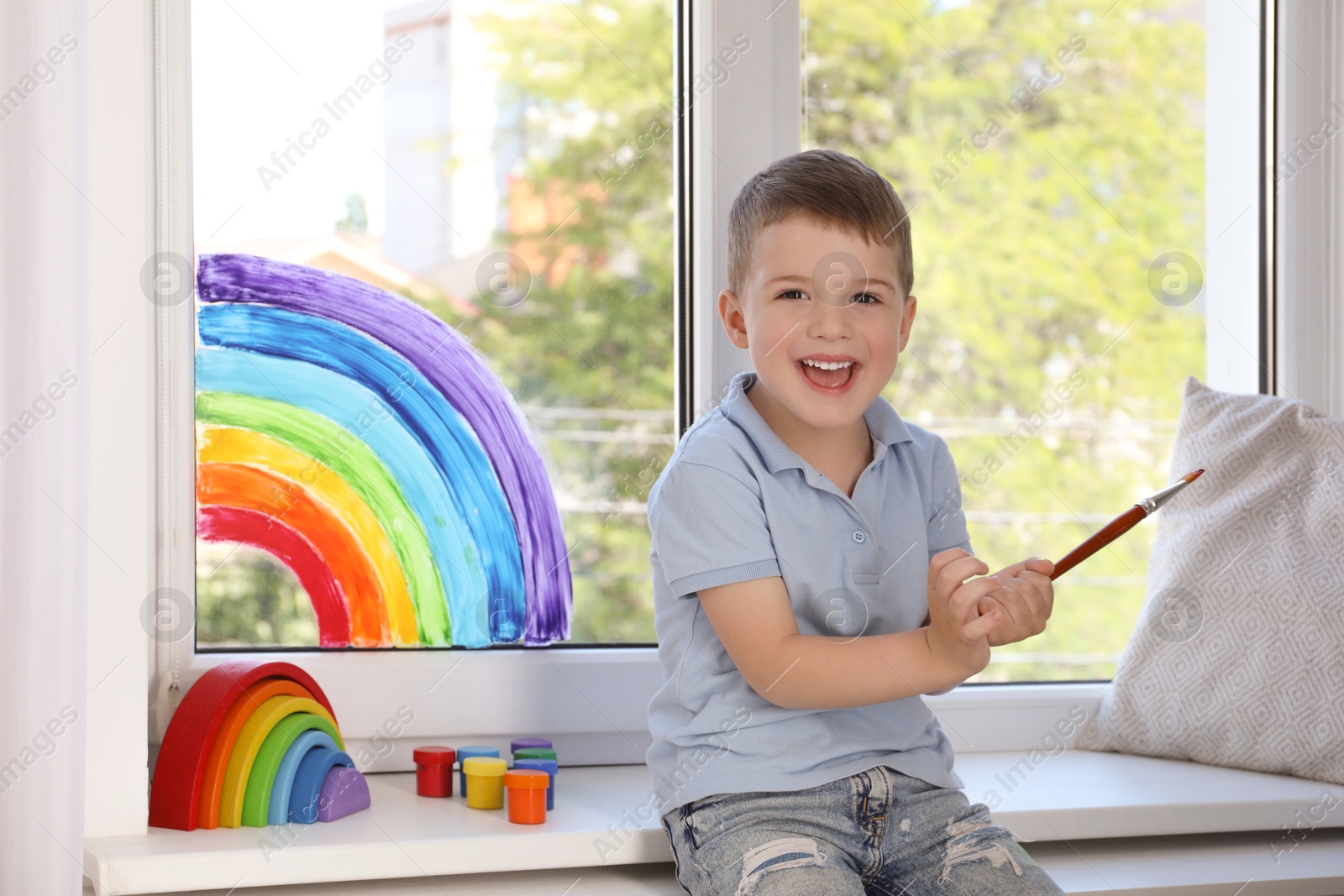 Photo of Little boy with brush near rainbow painting on window indoors