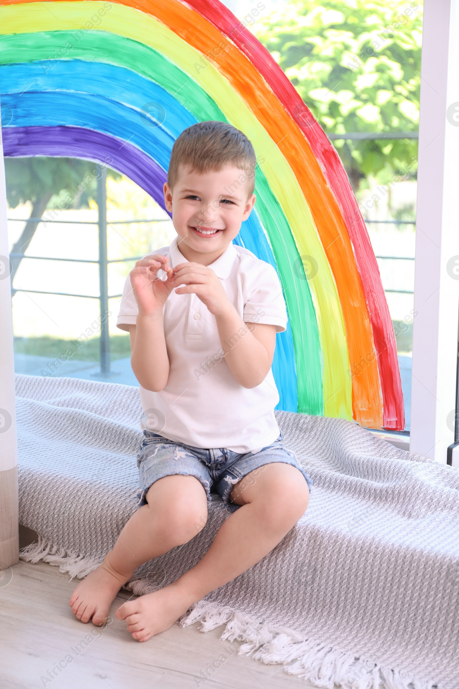 Photo of Little boy near rainbow painting on window indoors