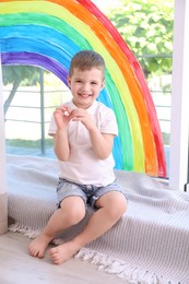 Photo of Little boy near rainbow painting on window indoors