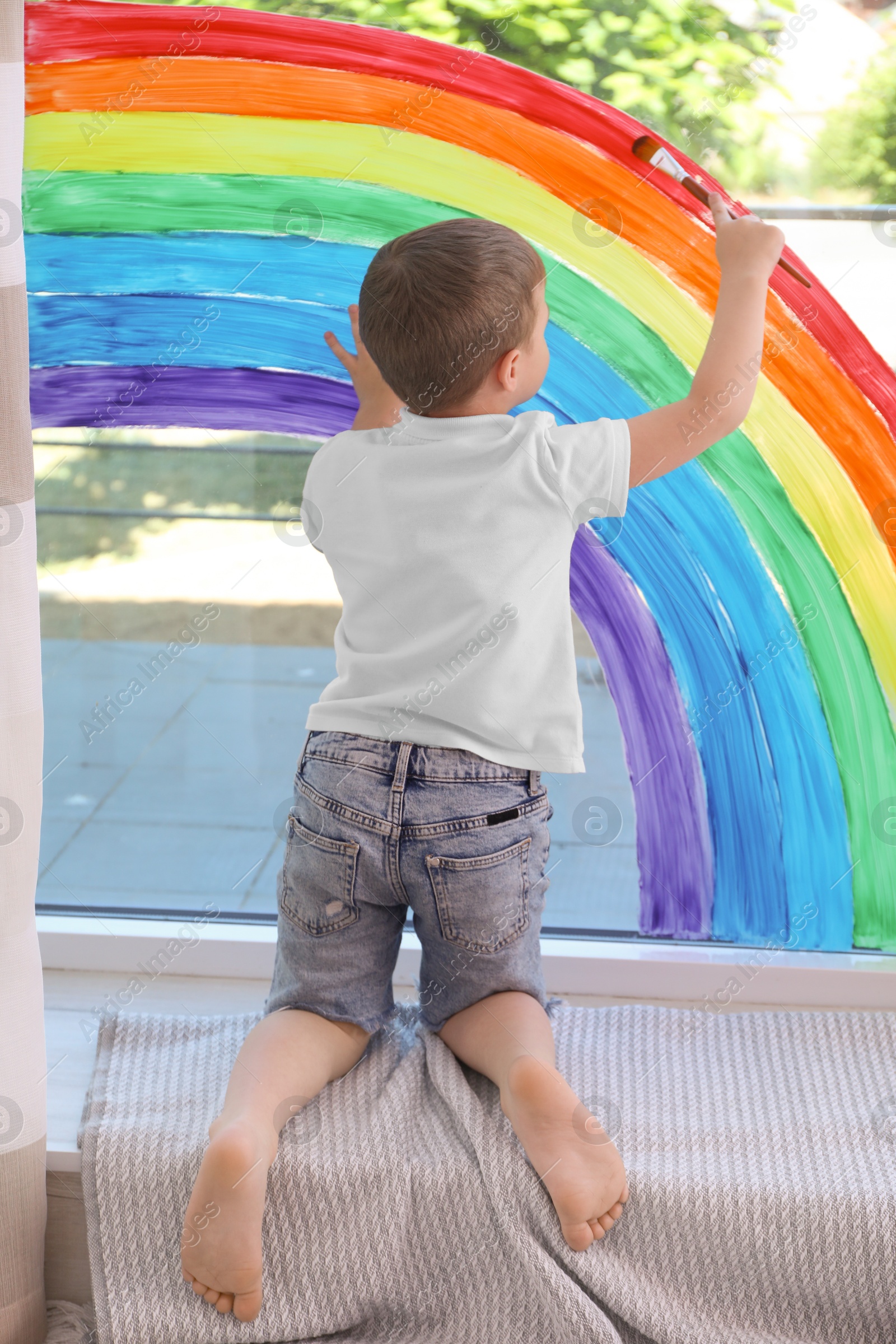 Photo of Little boy drawing rainbow on window indoors, back view