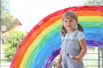 Photo of Little girl with brush near rainbow painting on window indoors