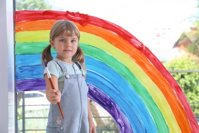 Photo of Little girl with brush near rainbow painting on window indoors
