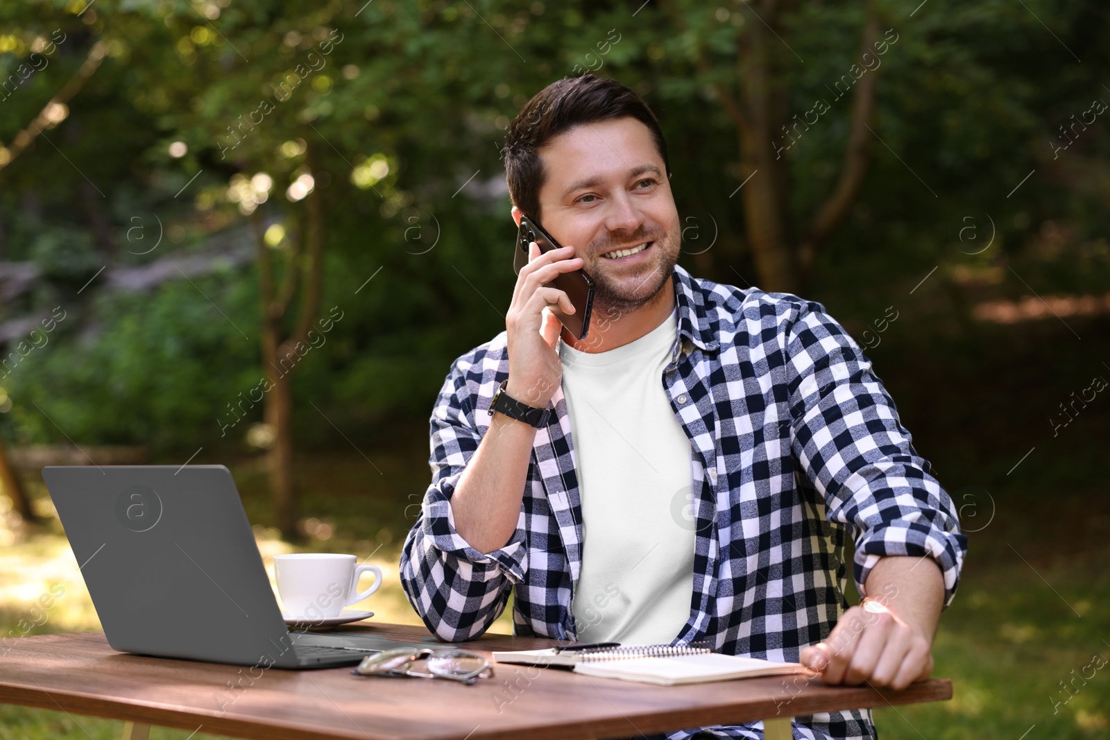 Photo of Smiling freelancer talking on smartphone at table with laptop outdoors. Remote job