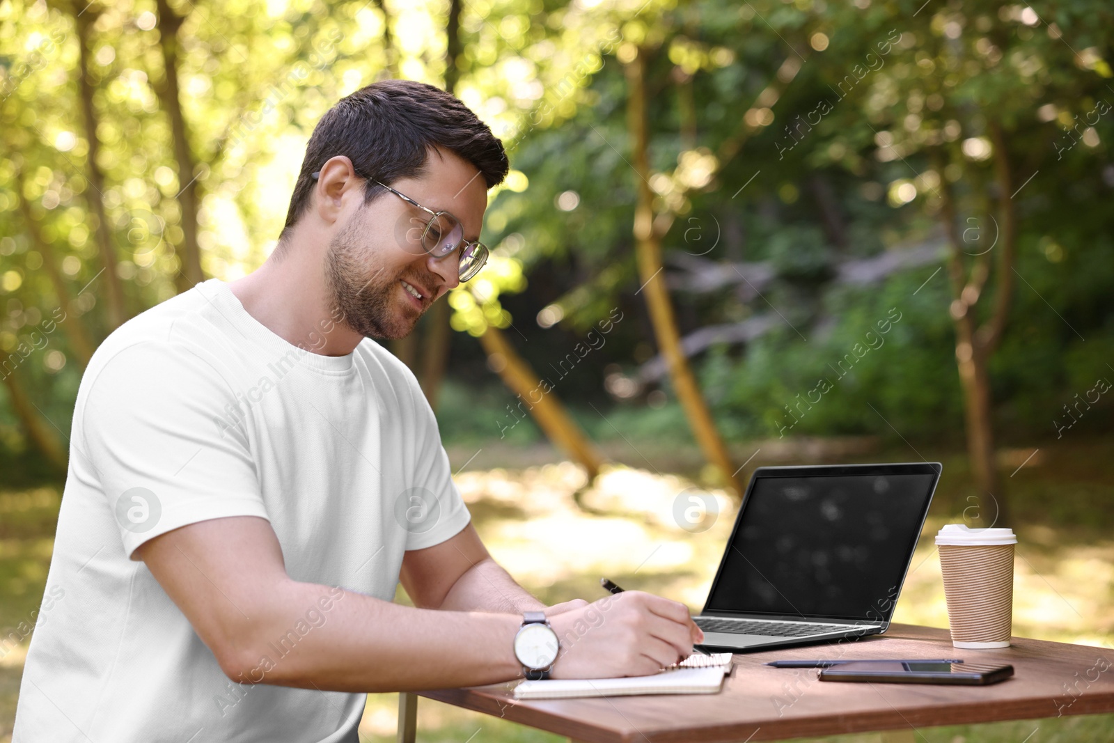 Photo of Smiling freelancer working with laptop and writing something at table outdoors. Remote job