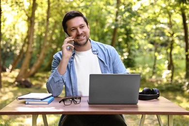 Smiling freelancer talking on smartphone at table with laptop outdoors. Remote job