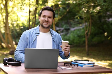 Smiling freelancer drinking coffee at table with laptop outdoors. Remote job