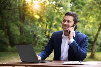 Smiling businessman talking on smartphone at table with laptop outdoors. Remote job