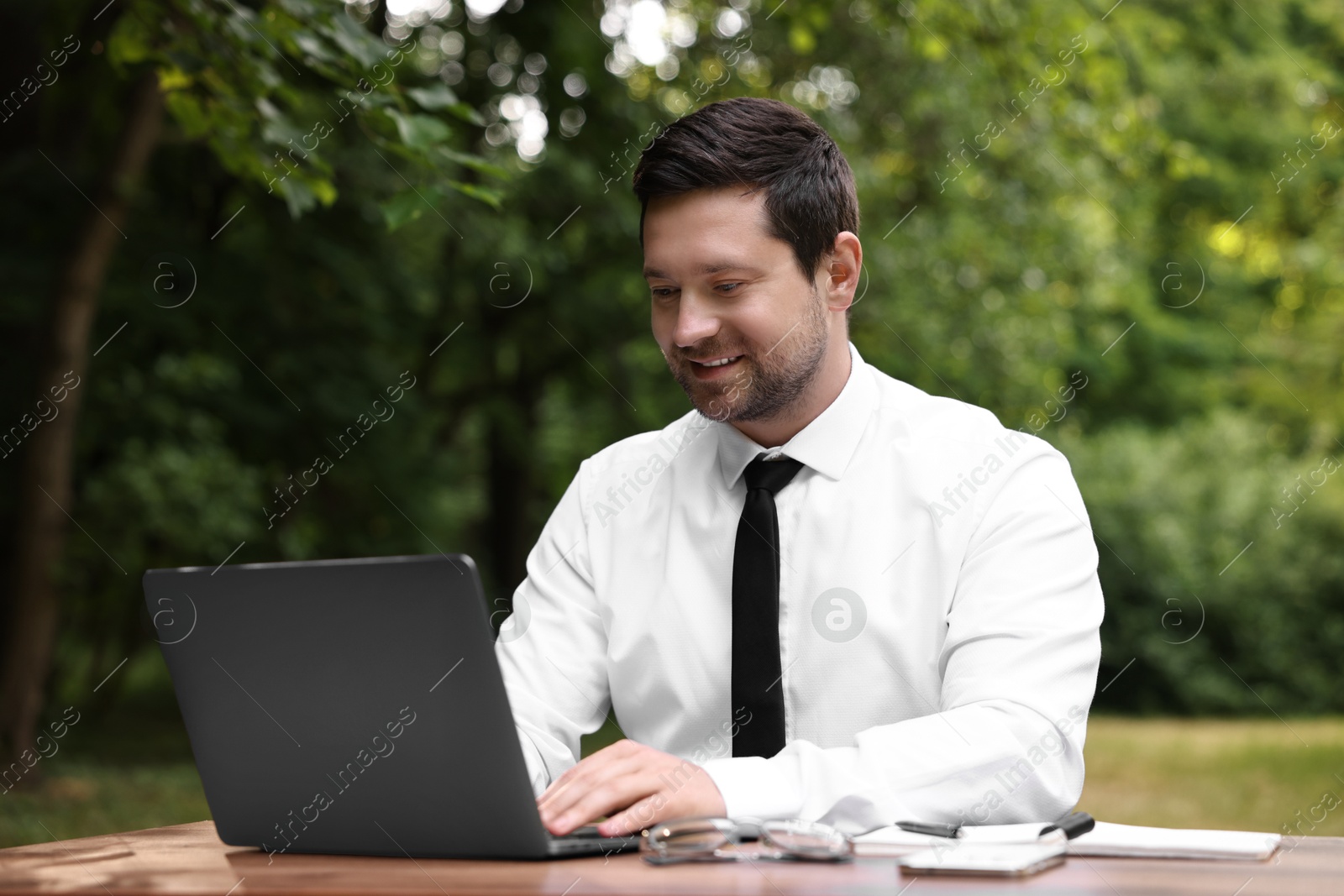 Photo of Smiling businessman working with laptop at table outdoors. Remote job