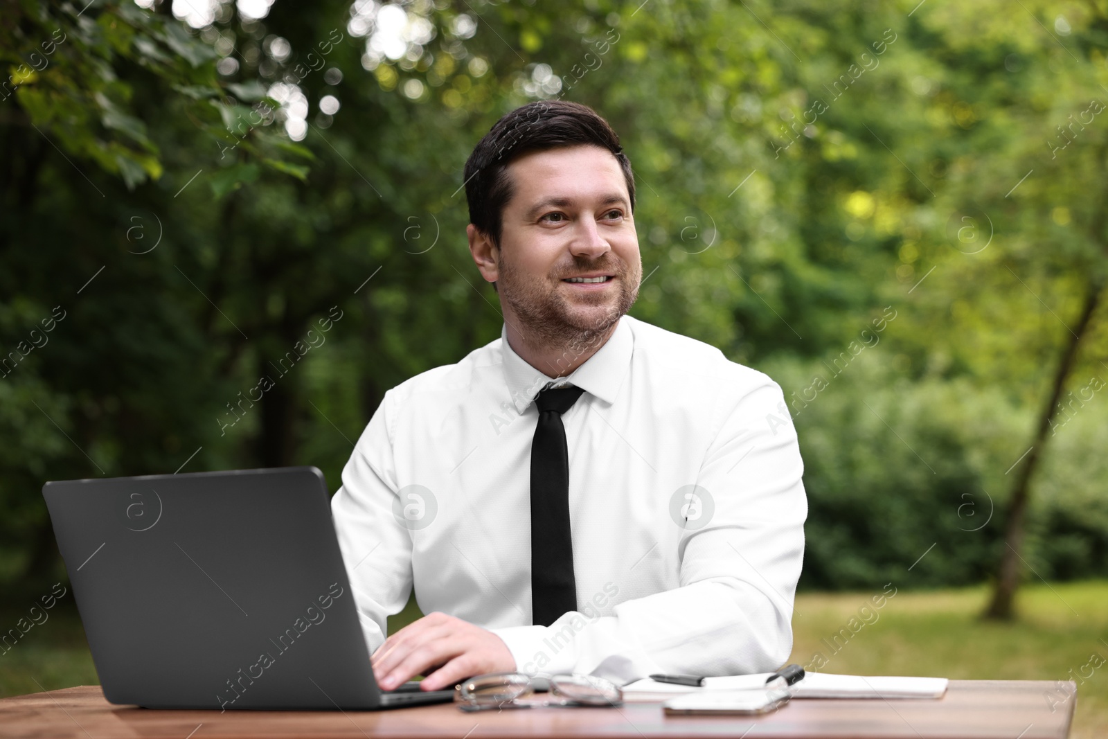 Photo of Smiling businessman working with laptop at table outdoors. Remote job