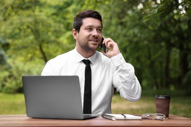 Businessman talking by smartphone at table with laptop outdoors. Remote job