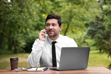 Photo of Businessman talking by smartphone at table with laptop outdoors. Remote job