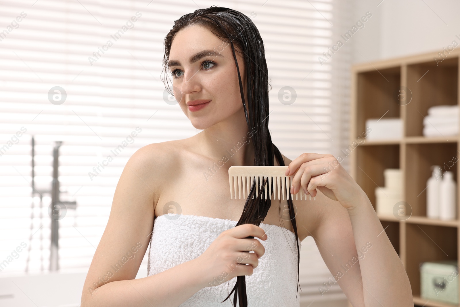 Photo of Beautiful woman combing her hair with applied mask in bathroom