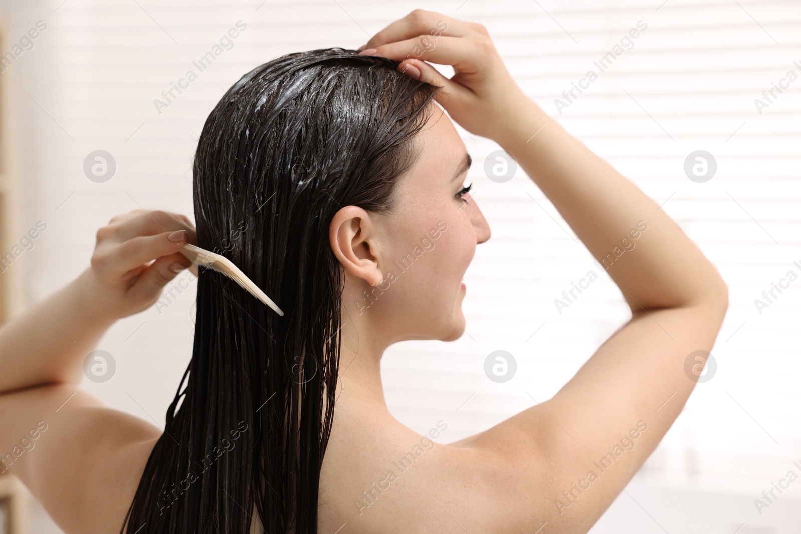 Photo of Woman combing her hair with applied mask in bathroom, back view