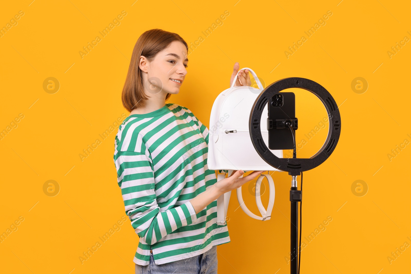 Photo of Fashion blogger reviewing backpack and recording video with smartphone and ring lamp on orange background