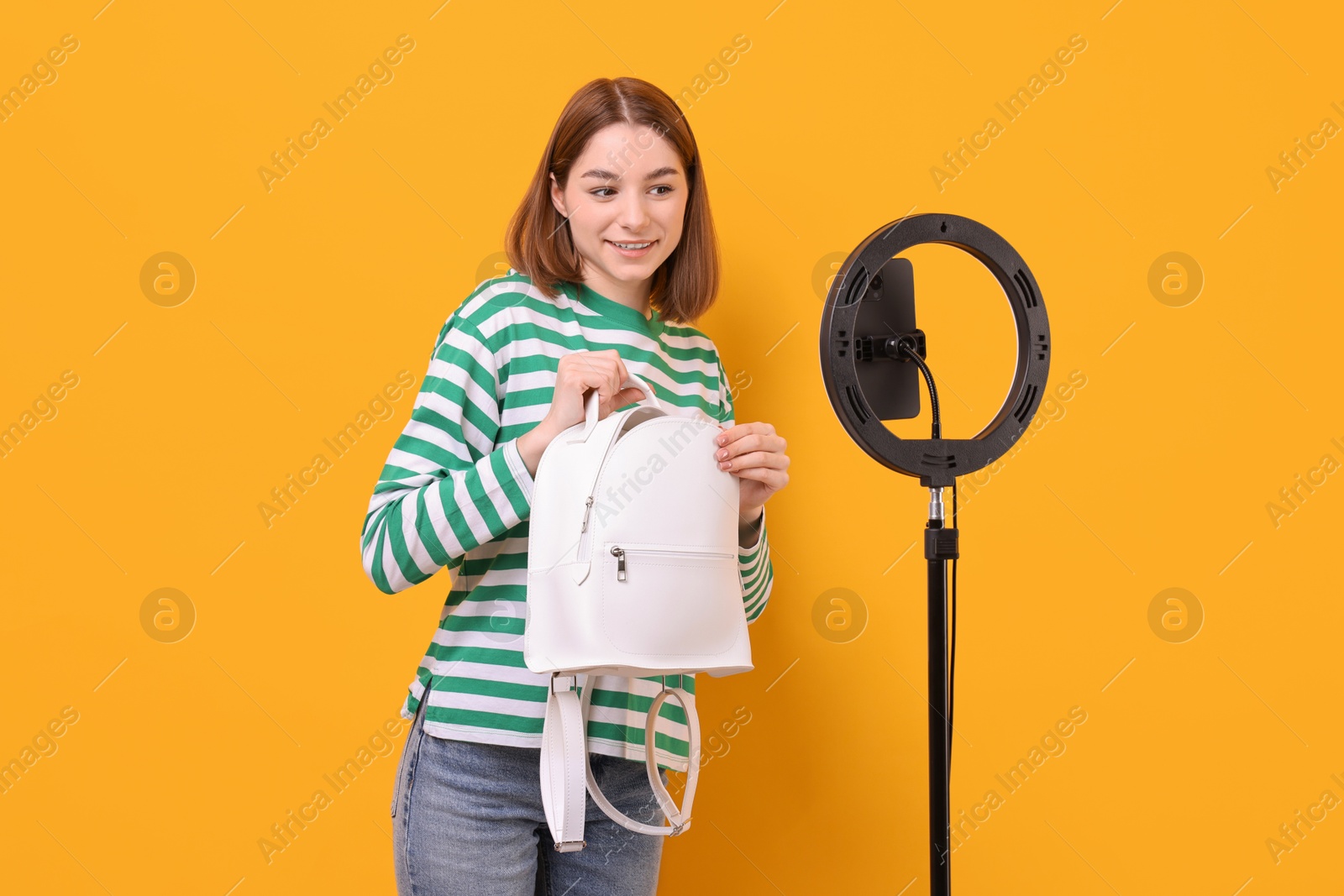 Photo of Fashion blogger reviewing backpack and recording video with smartphone and ring lamp on orange background