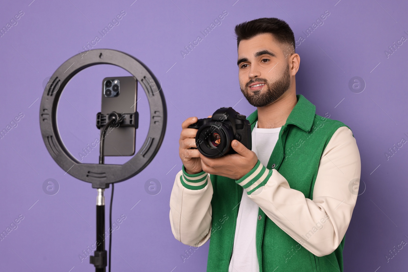 Photo of Technology blogger reviewing camera and recording video with smartphone and ring lamp on purple background