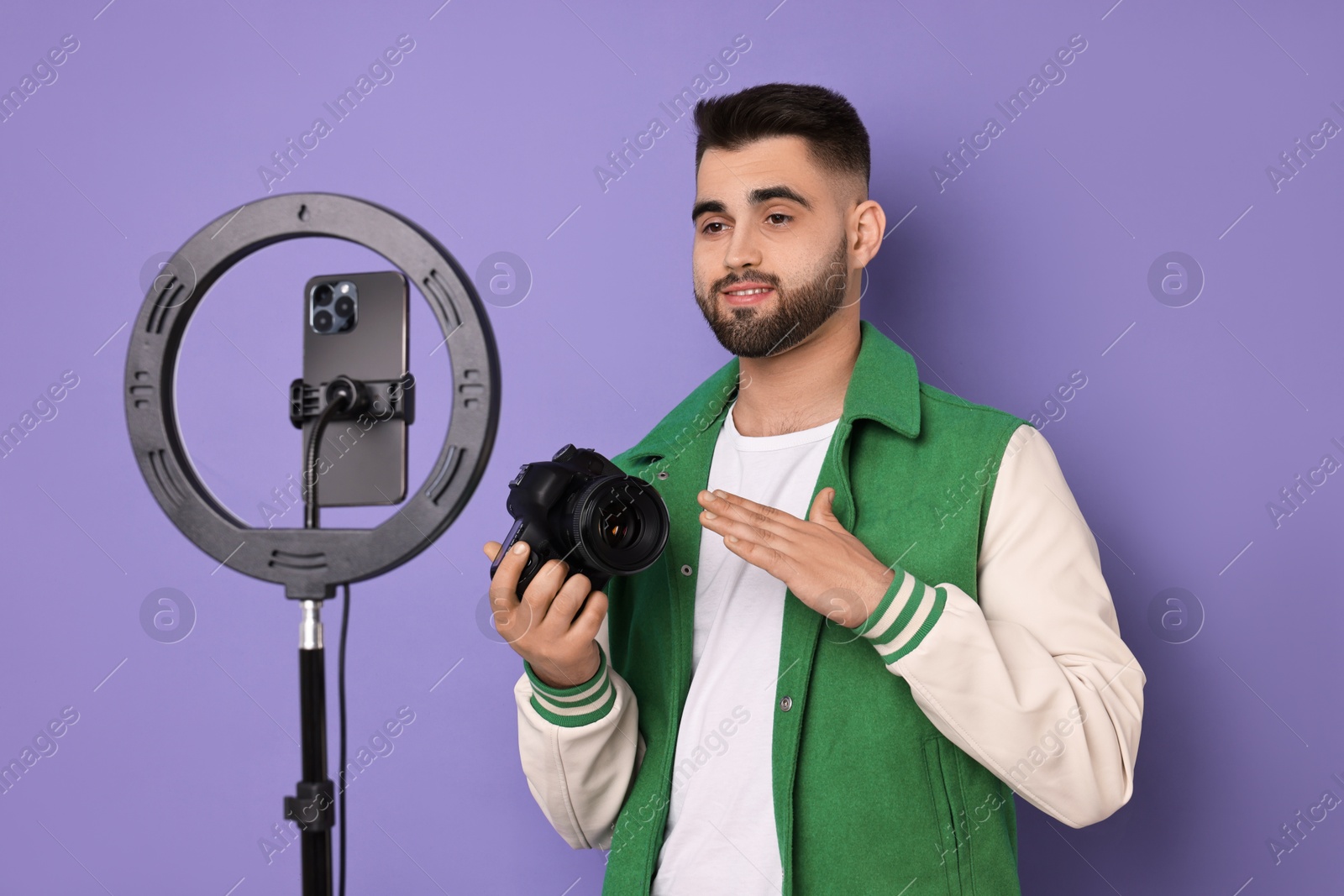 Photo of Technology blogger reviewing camera and recording video with smartphone and ring lamp on purple background