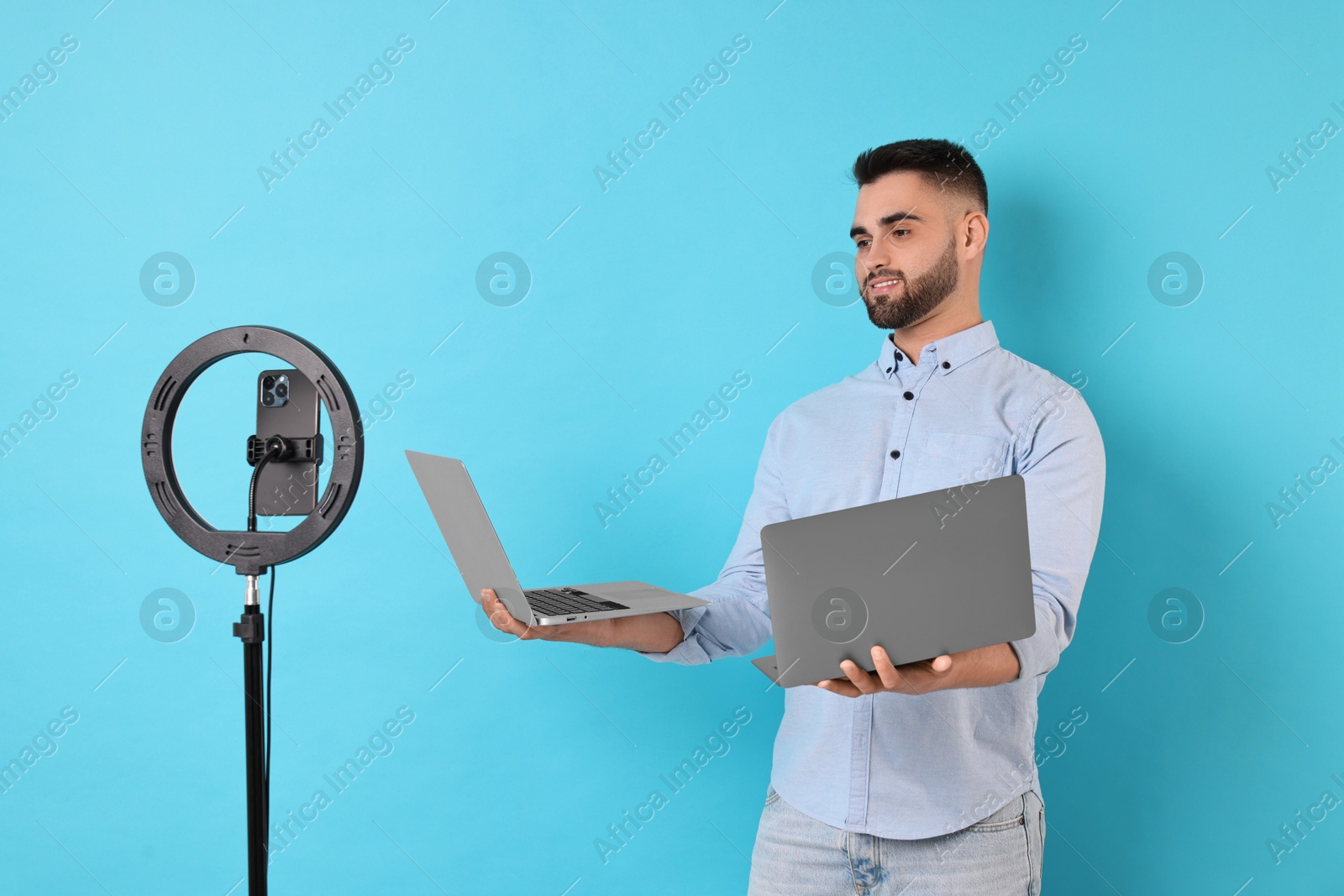 Photo of Technology blogger reviewing laptops and recording video with smartphone and ring lamp on light blue background