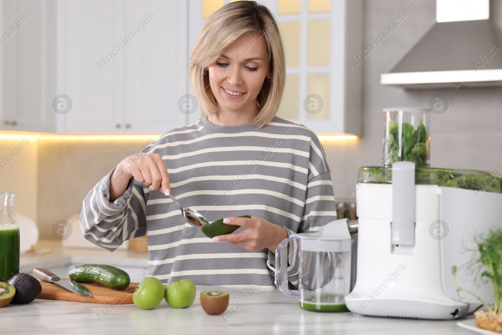 Photo of Smiling woman with fresh products using juicer at white marble table in kitchen