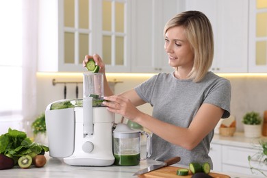 Photo of Smiling woman putting fresh cucumber into juicer at white marble table in kitchen