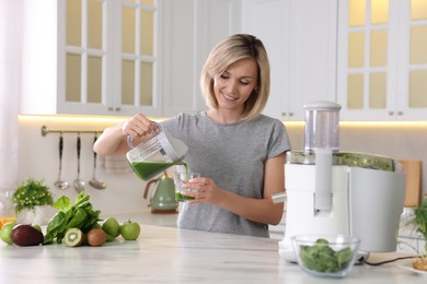 Smiling woman pouring juice into glass in kitchen. Juicer and fresh products on white marble table