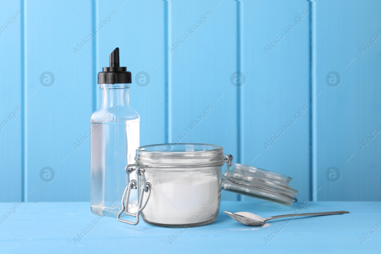 Photo of Natural cleaning products. Vinegar in bottle, baking soda and spoon on light blue wooden table