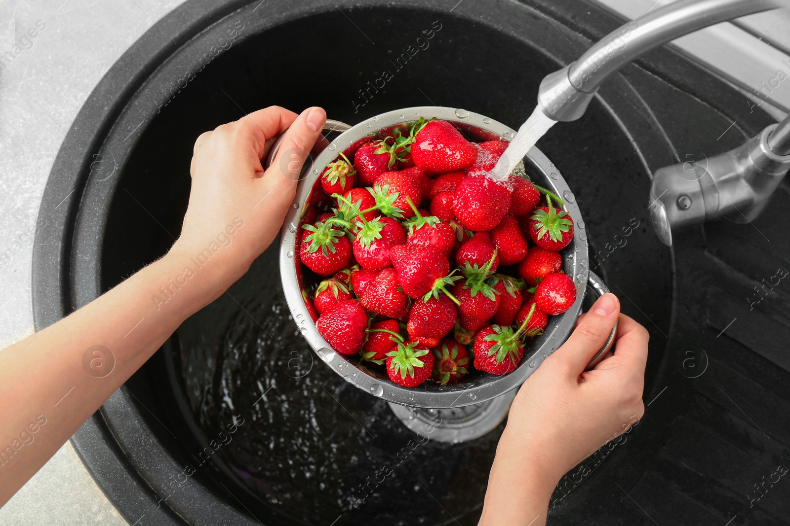 Photo of Woman washing fresh strawberries under tap water in metal colander above sink, top view