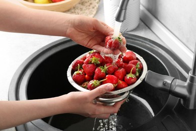 Photo of Woman washing fresh strawberries under tap water in metal colander above sink, closeup