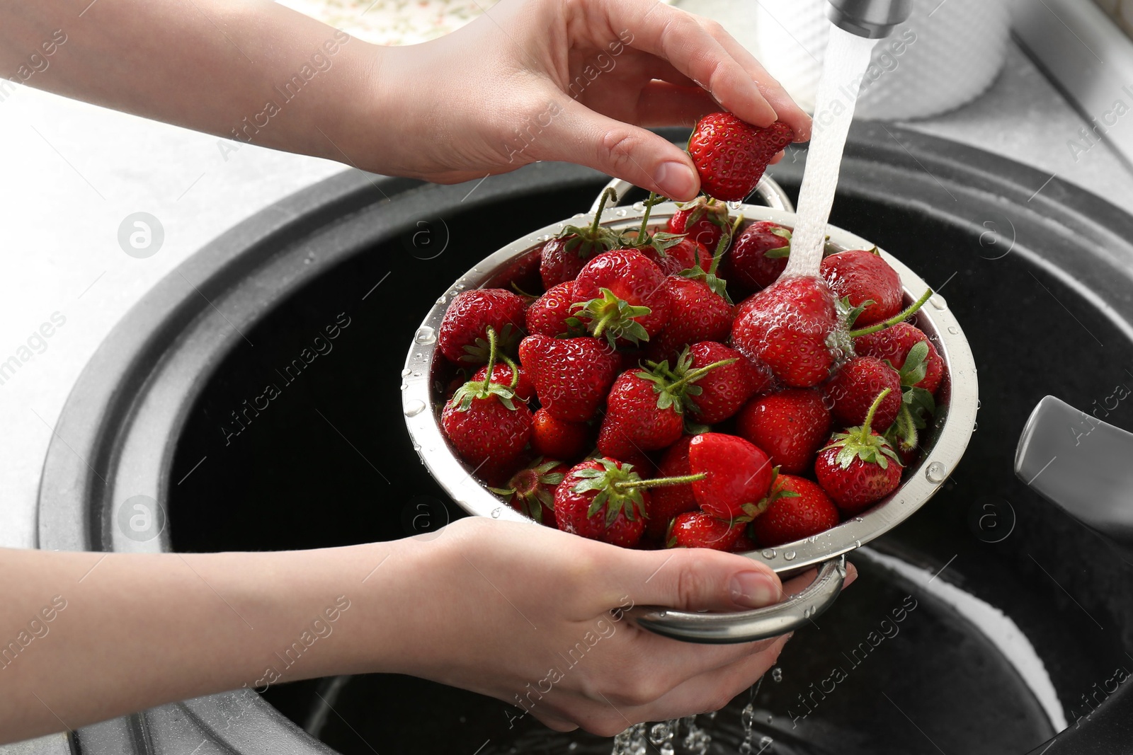 Photo of Woman washing fresh strawberries under tap water in metal colander above sink, closeup