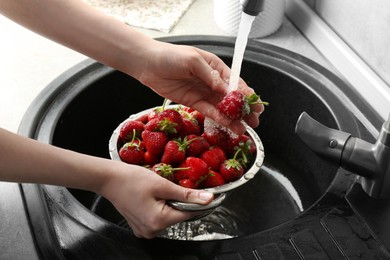 Photo of Woman washing fresh strawberries under tap water in metal colander above sink, closeup