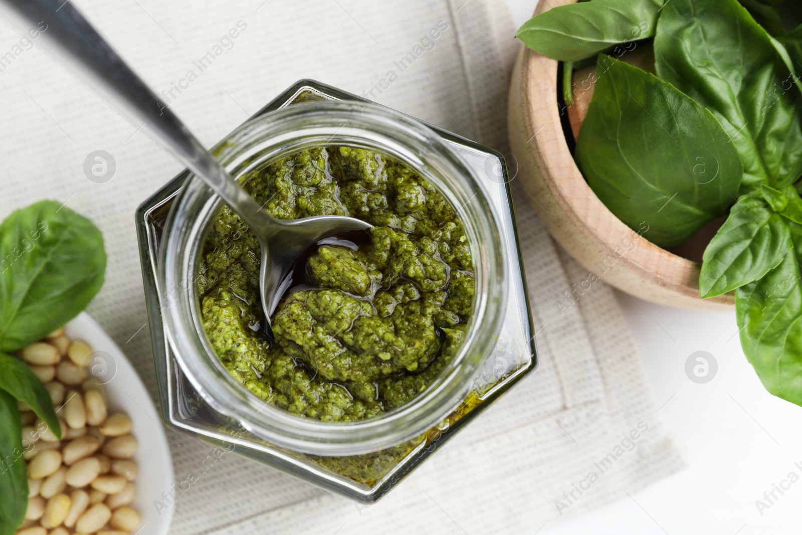 Photo of Tasty pesto sauce in glass jar, spoon, pine nuts and basil on white table, top view