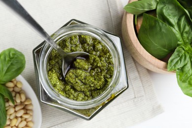 Tasty pesto sauce in glass jar, spoon, pine nuts and basil on white table, top view