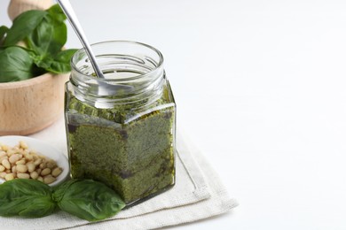 Photo of Tasty pesto sauce in glass jar, spoon, pine nuts and basil on white table, space for text