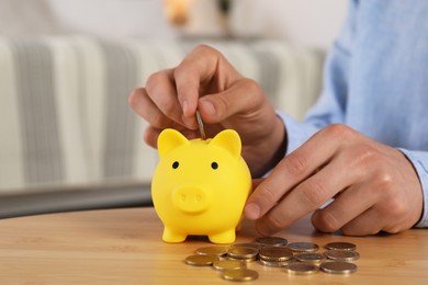 Photo of Man putting coin into yellow piggy bank on wooden table, closeup. Space for text