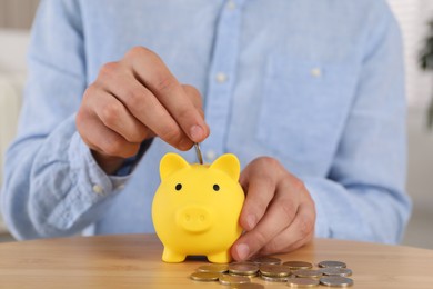 Photo of Man putting coin into yellow piggy bank on wooden table, closeup