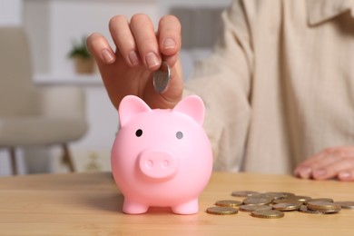 Photo of Woman putting coin into pink piggy bank at wooden table, closeup