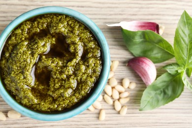 Photo of Tasty pesto sauce in bowl, pine nuts, garlic and basil on wooden table, top view