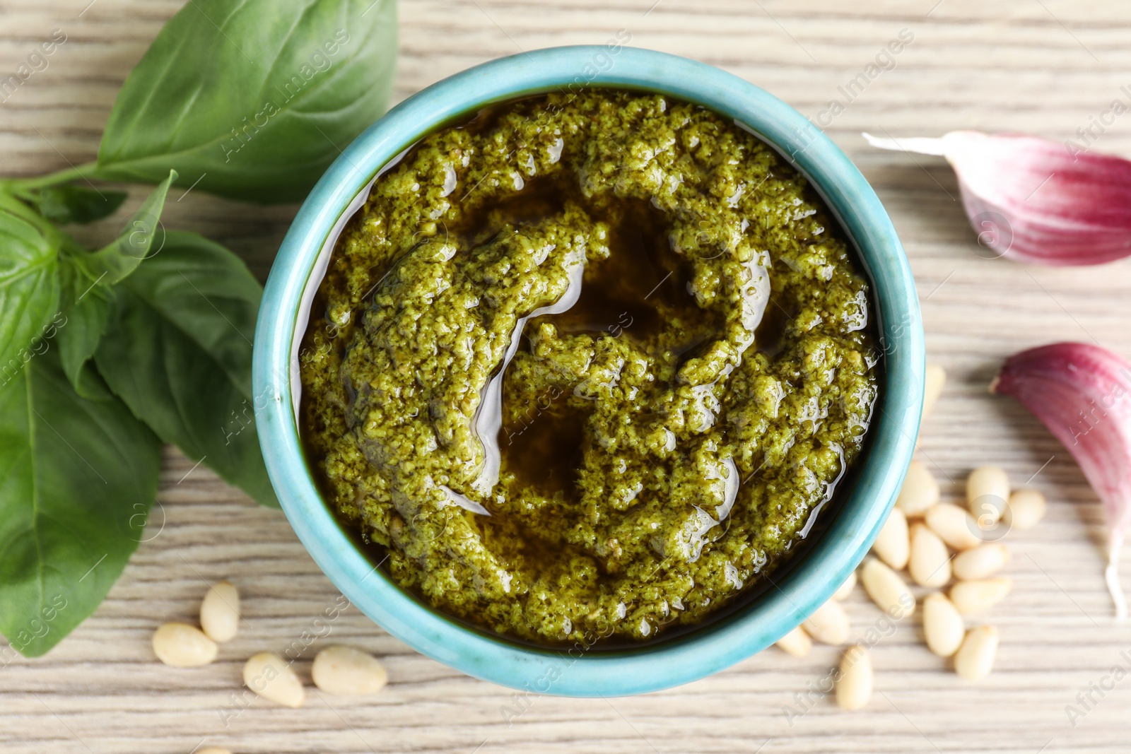 Photo of Tasty pesto sauce in bowl, pine nuts, garlic and basil on wooden table, top view