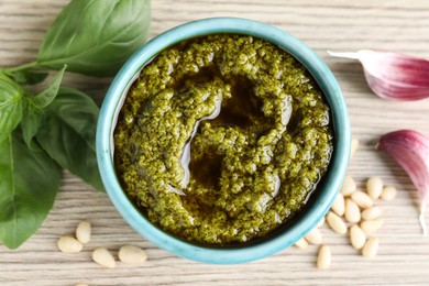 Photo of Tasty pesto sauce in bowl, pine nuts, garlic and basil on wooden table, top view