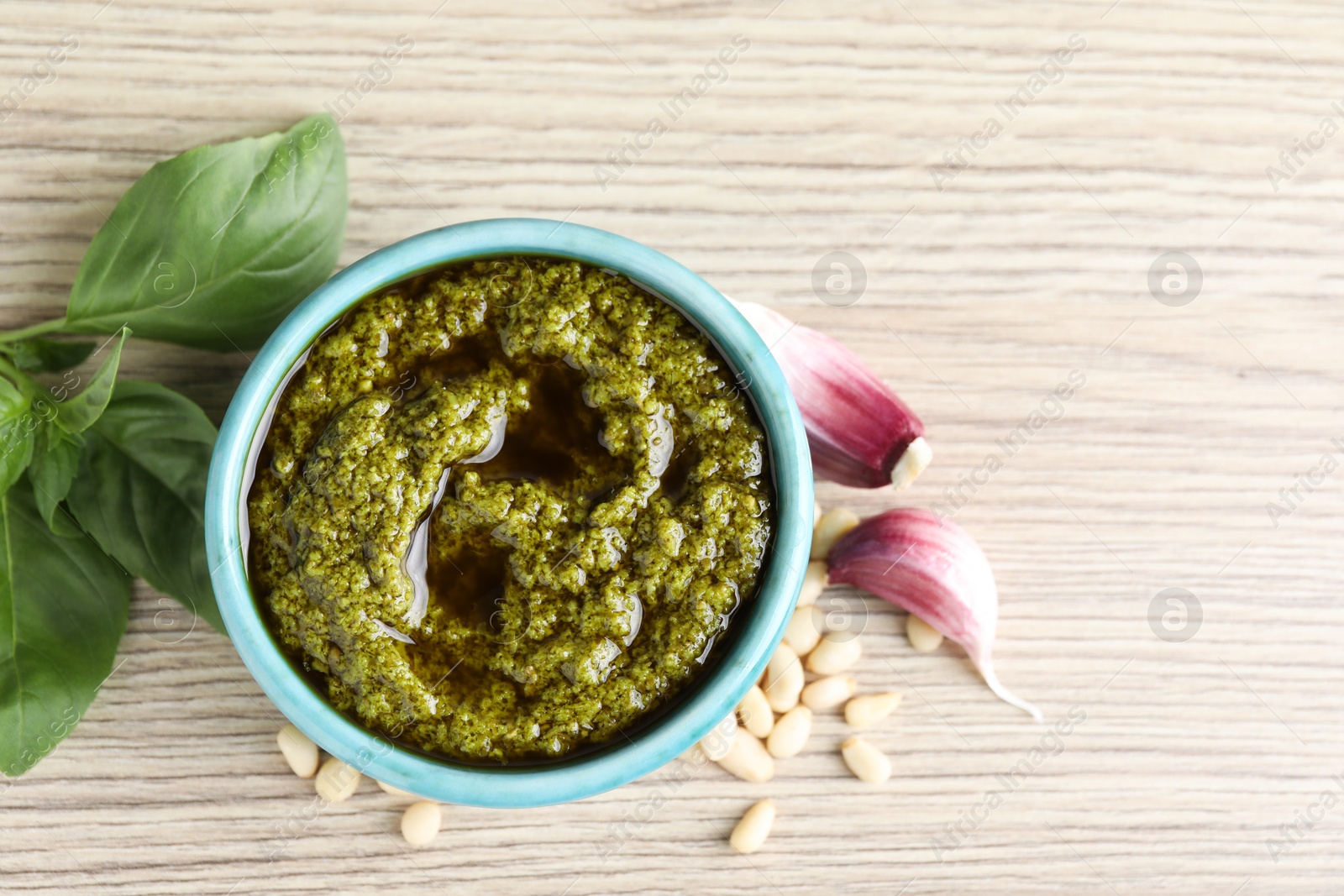 Photo of Tasty pesto sauce in bowl, pine nuts, garlic and basil on wooden table, top view. Space for text