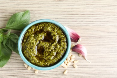 Tasty pesto sauce in bowl, pine nuts, garlic and basil on wooden table, top view. Space for text