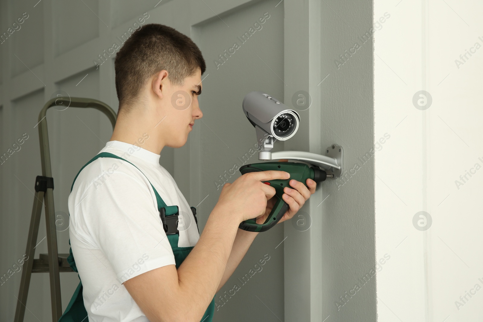 Photo of Technician with cordless electric screwdriver installing CCTV camera on wall indoors