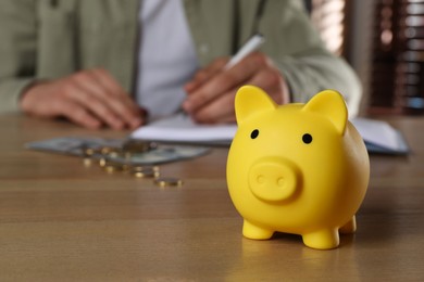 Photo of Man at wooden table, focus on yellow piggy bank. Space for text